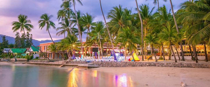 Strand bei Sonnenuntergang mit bewölktem Himmel; zahlreiche Palmen im Fokus, ein beleuchtetes Hotel im Hintergrund. Bunte Deko spiegelt sich im Wasser und sorgt für eine farbenfrohe Atmosphäre.